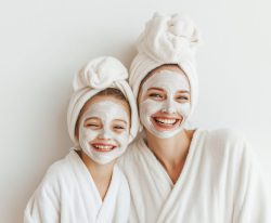 Teenage girl and her mom apply a clay white skin care mask to her face. They are in robes and with a towel on their heads at the spa. White cosmetics or beauty salon banner background with copy space.